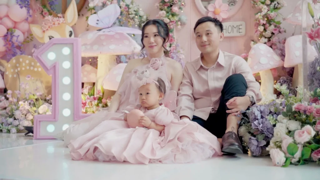 Anak perempuan berpakaian pink, duduk di antara orang tua di depan dekorasi tema pink.