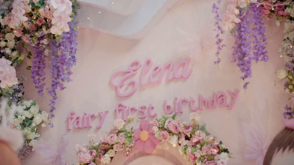 Backdrop perayaan ulang tahun Elena dengan bunga dan huruf "Elena fairy first birthday".