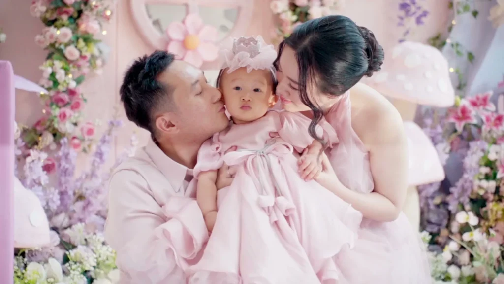Orang tua memeluk bayi perempuan yang mengenakan gaun pink, latar belakang berwarna pink dan bunga.
