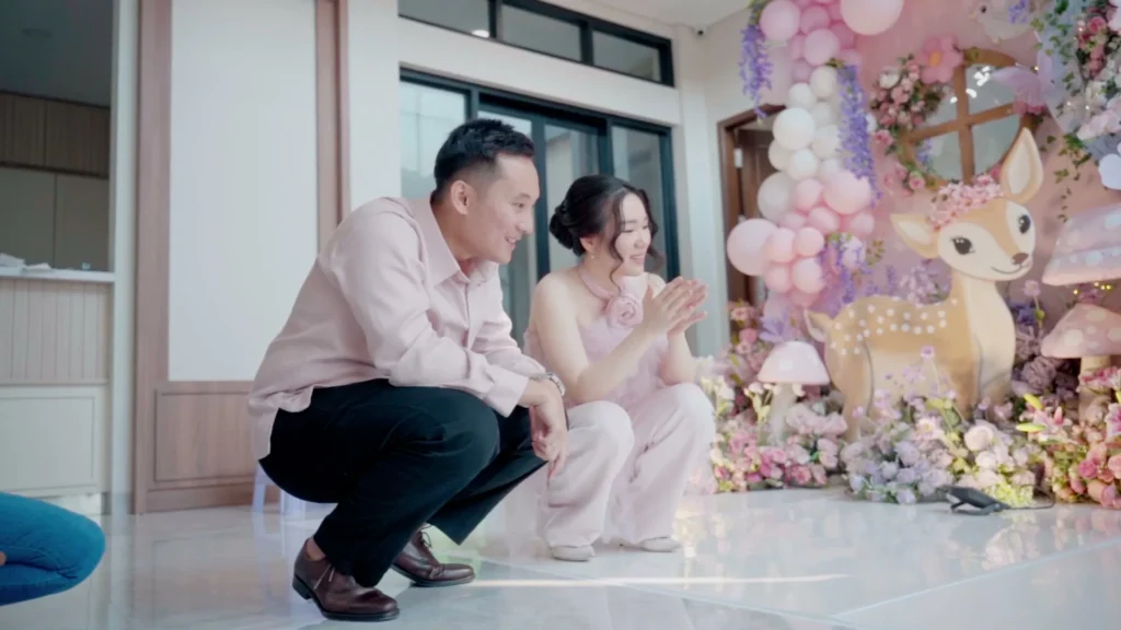 Pasangan berpakaian formal berpose di depan dekorasi ulang tahun dengan tema dekorasi berwarna pastel.